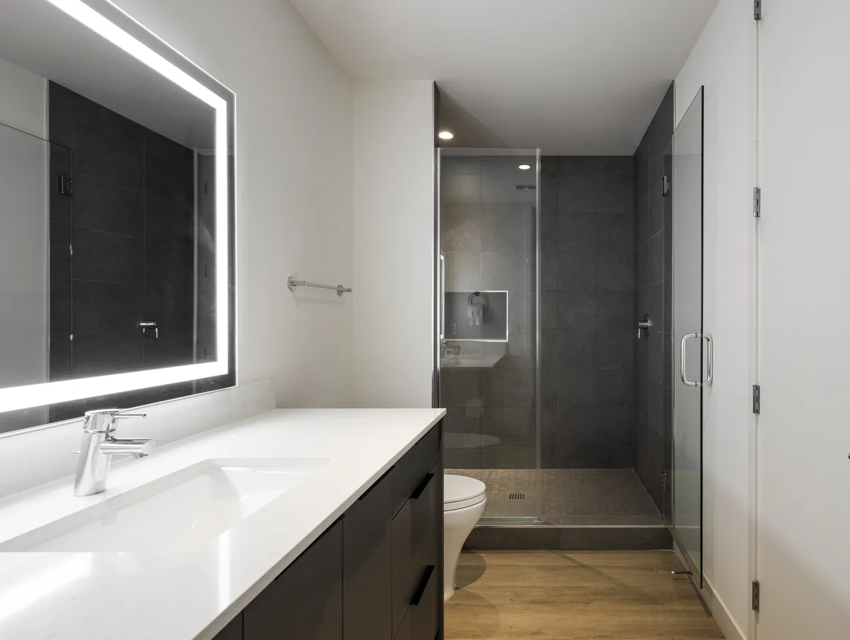 This modern bathroom features a dual sink vanity with illuminated mirrors and a walk-in glass shower enclosure. The clean lines and bright, serene lighting evoke a feeling of sophistication and calm, perfect for refreshing after a long day.