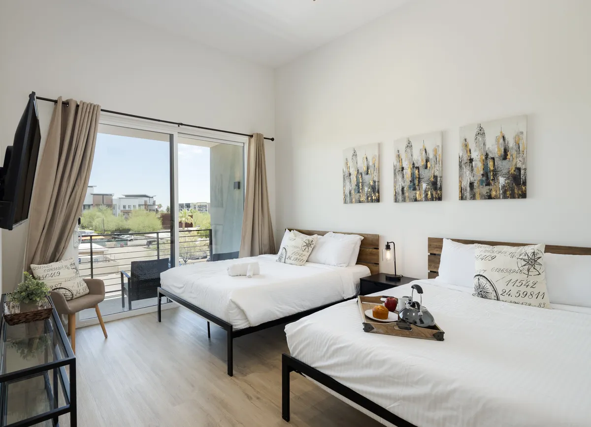 This image shows an inviting bedroom suite featuring two queen beds with crisp white linens, stylish decorative artwork on the wall, and a breakfast tray resting atop the bed. Floor-to-ceiling windows open onto a private balcony, allowing natural light to fill the space and offering serene outdoor views.