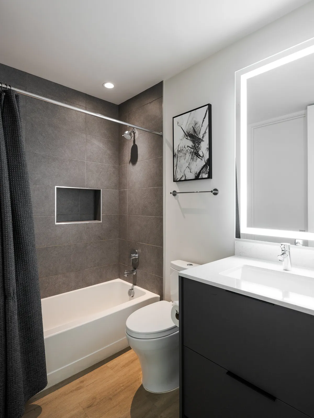 A contemporary en-suite bathroom featuring a sleek soaking tub, illuminated vanity mirror, subtle wall art, and an elegant vanity unit. The space is bathed in soft ambient light, evoking a spa-like atmosphere ideal for relaxation and self-care.