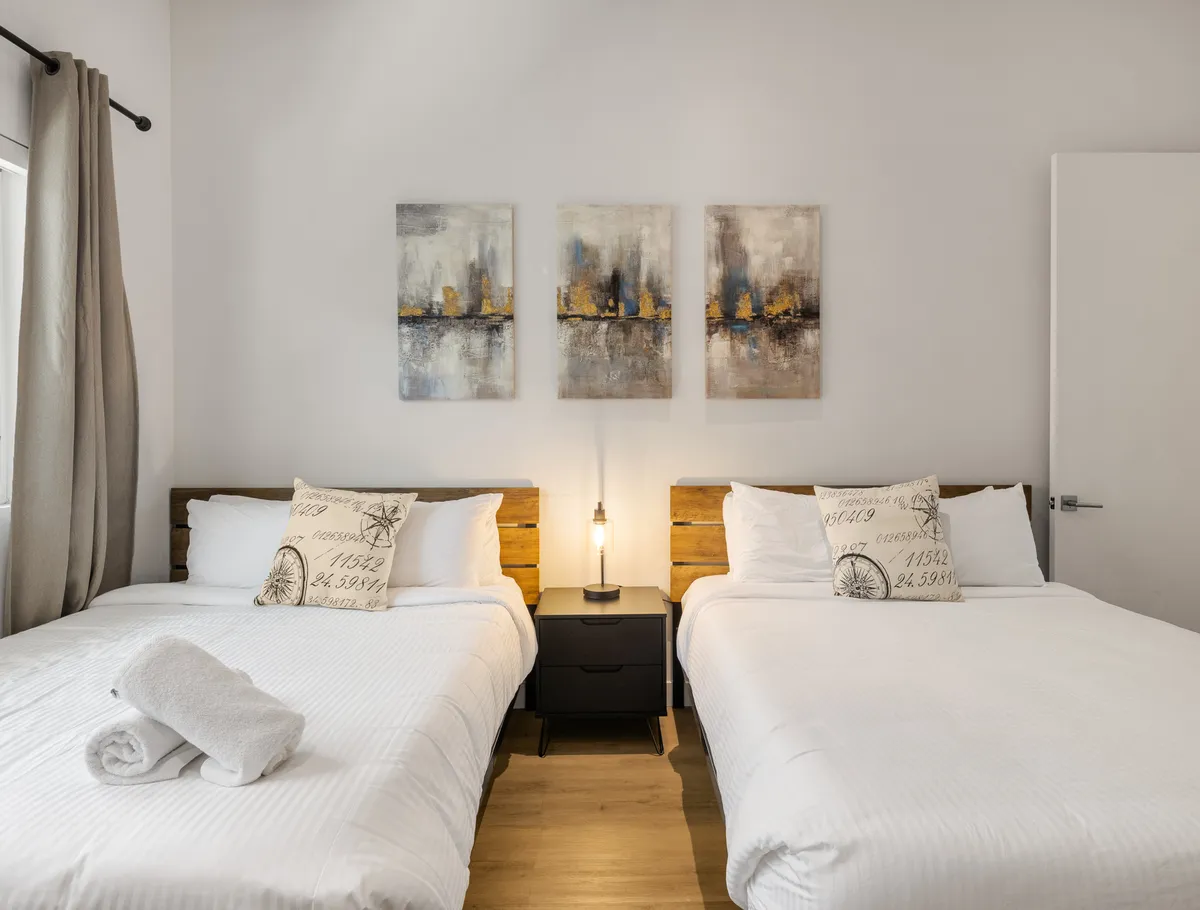 The photo centers on a tranquil bedroom arrangement with two queen beds dressed in soft white linens, tasteful artwork above the beds, and a shared nightstand with an elegant lamp. The cozy, uncluttered design conveys a restful atmosphere ideal for families or friends.