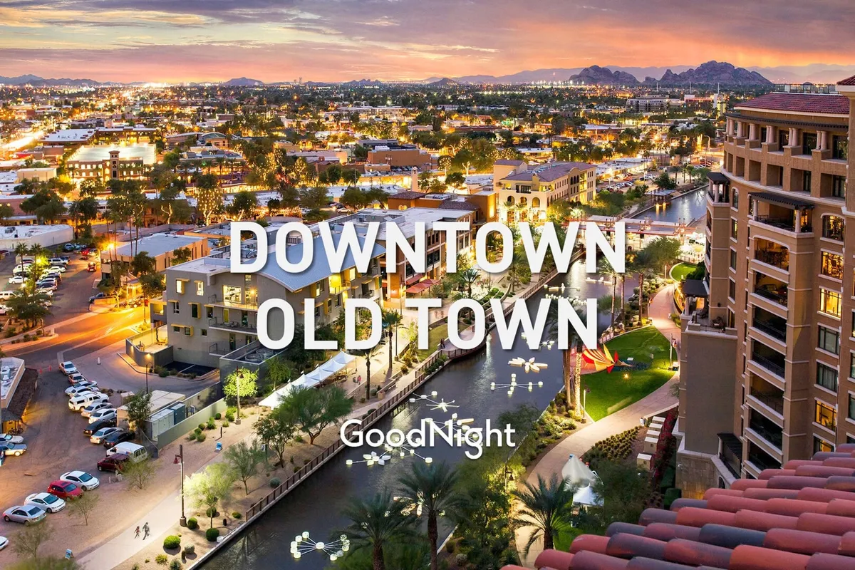 A lively nighttime scene with illuminated buildings, lively canal reflections, palm trees, and people dining al fresco alongside the water. The mountains serve as a distant backdrop, and the area is bustling with nightlife activity and outdoor restaurants.