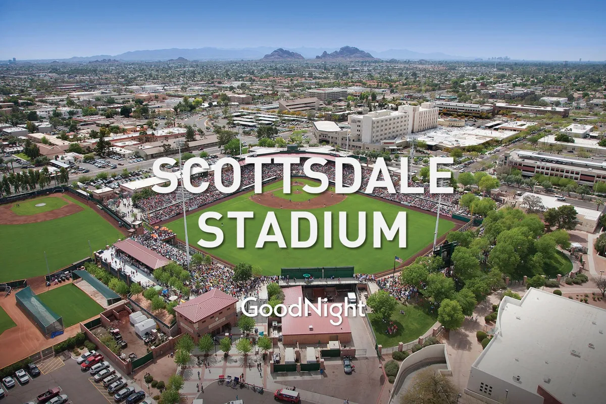 An exterior shot of a bustling baseball stadium with a crowd enjoying a lively game, flanked by surrounding buildings and a mountain backdrop. The communal, energetic atmosphere is palpable.