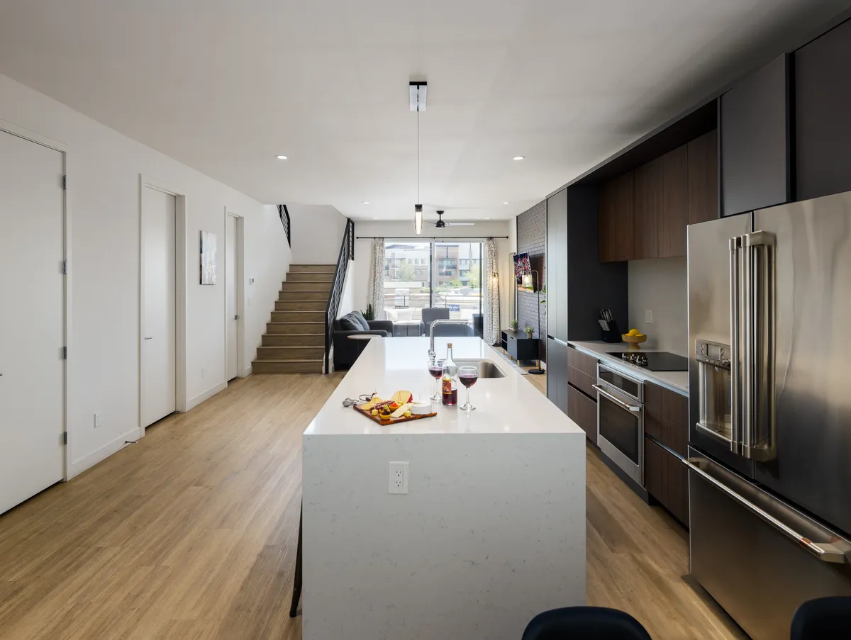 This image features a modern interior with a visible staircase, kitchen island, stainless steel appliances, large window, and living area. The staircase is a prominent element in the space, highlighting the multi-level layout of the property.