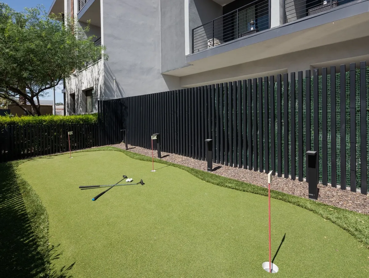 An outdoor putting green bordered by a manicured green hedge, with golf clubs set up and modern architecture visible in the background. The area is fenced for privacy and offers an inviting spot for golf practice or relaxed outdoor fun.