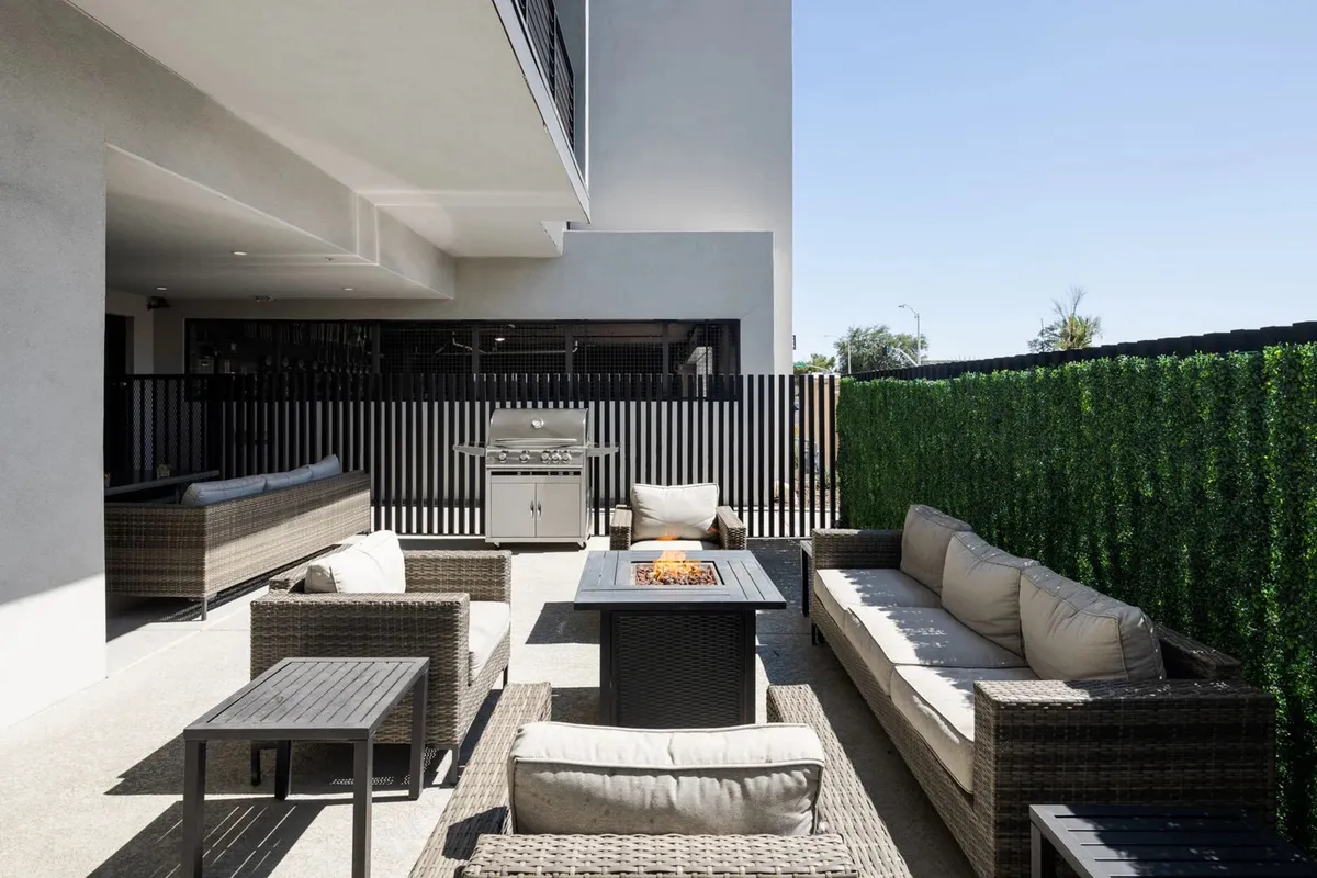 A cozy outdoor lounge area centered around a built-in fire pit, with light-colored cushioned seating, a BBQ grill to one side, and lush green hedges providing privacy. The space is designed for group gatherings, alfresco dining, and evening relaxation under the stars.