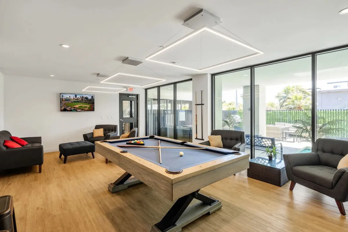 A stylish communal clubhouse featuring a pool table at the center, surrounded by comfortable seating and large windows that let in plenty of natural light. A mounted TV screen provides a focal point for watching sports or movies, while the overall atmosphere is casual and welcoming, inviting guests to relax or engage in friendly competition.
